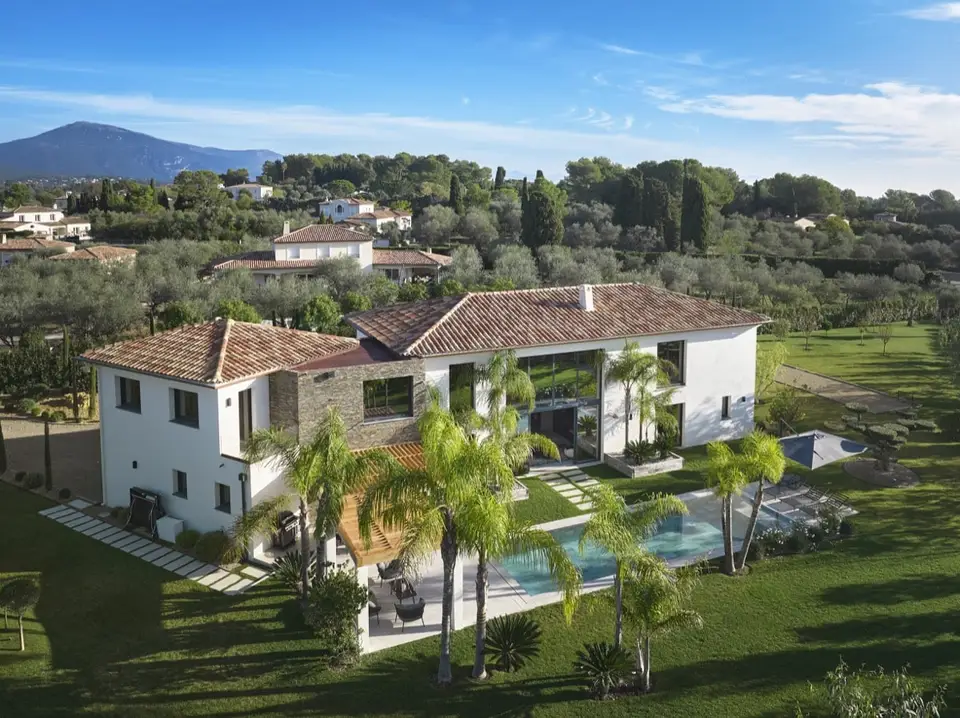 Aerial view of a villa with tiled roofs surrounded by palm trees, olive trees and lawn, with the pool and the hills in the background.