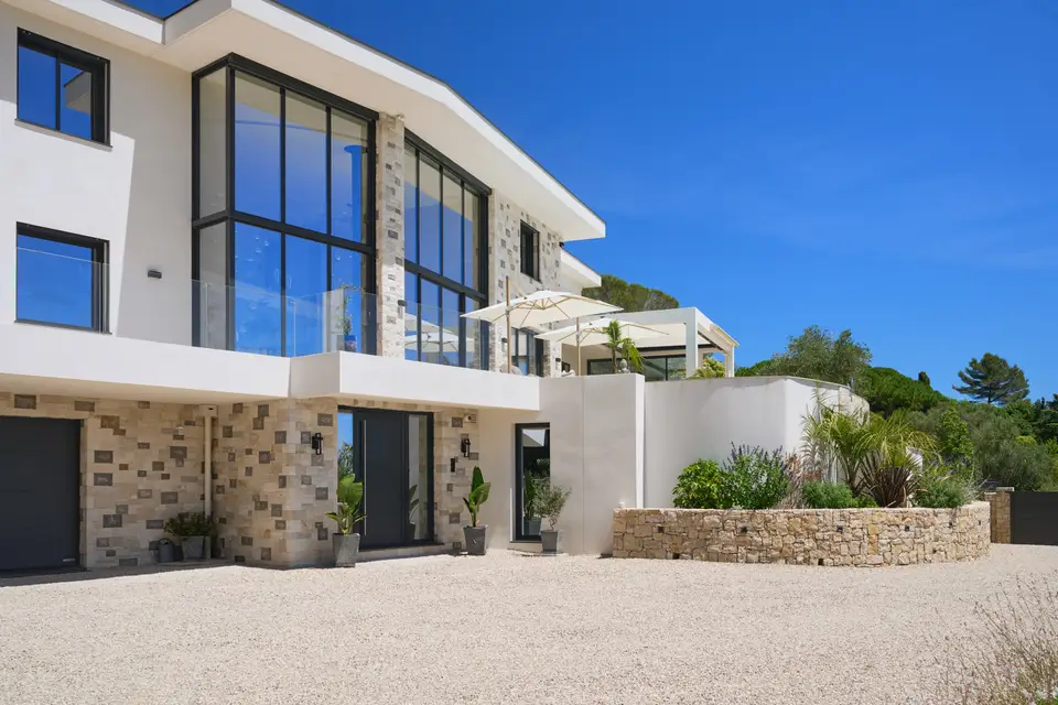 Entrance façade of a white contemporary villa with a large black-framed glazed wall, pale stone cladding and wide roof overhangs.