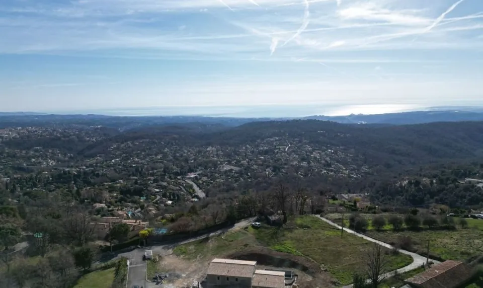 Drone view over the hills of Tourrettes-sur-Loup: the plot and its access track in the foreground, wooded hills and scattered houses down to the distant sea