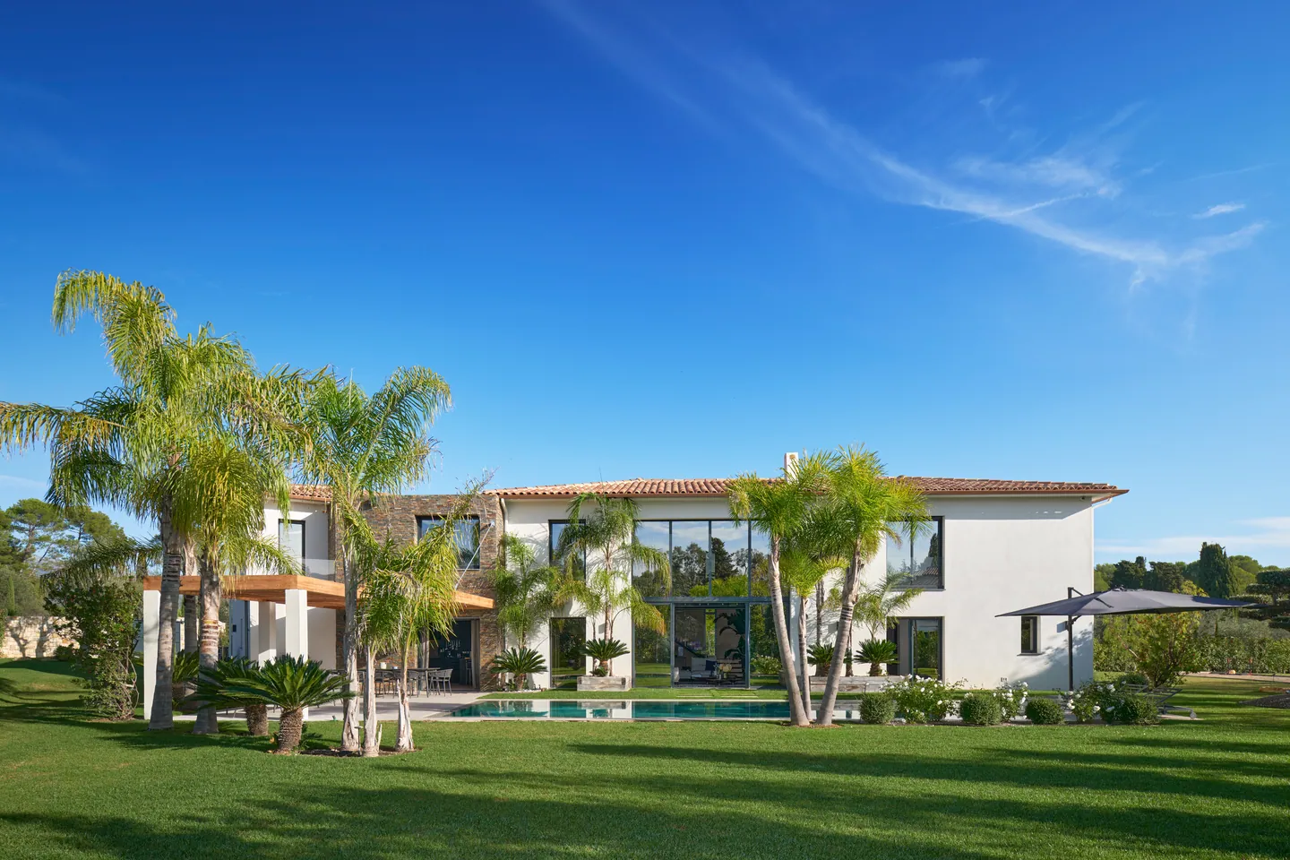 Façade côté jardin avec grande baie vitrée, palmiers et piscine au bout de la pelouse