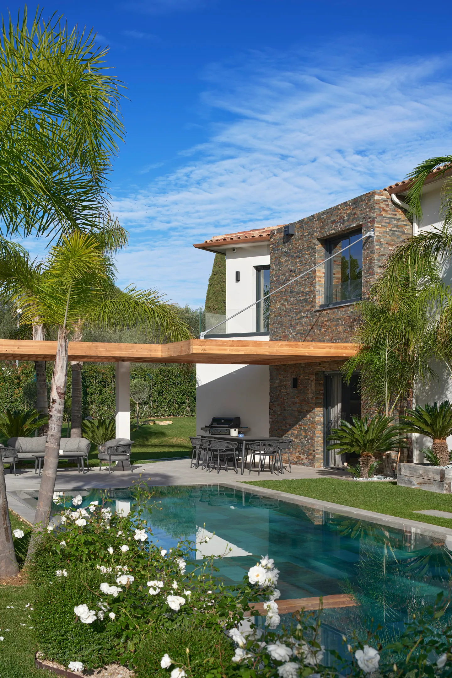 Piscine bordée de rosiers blancs, pergola en bois et repas d'été devant le volume en pierre