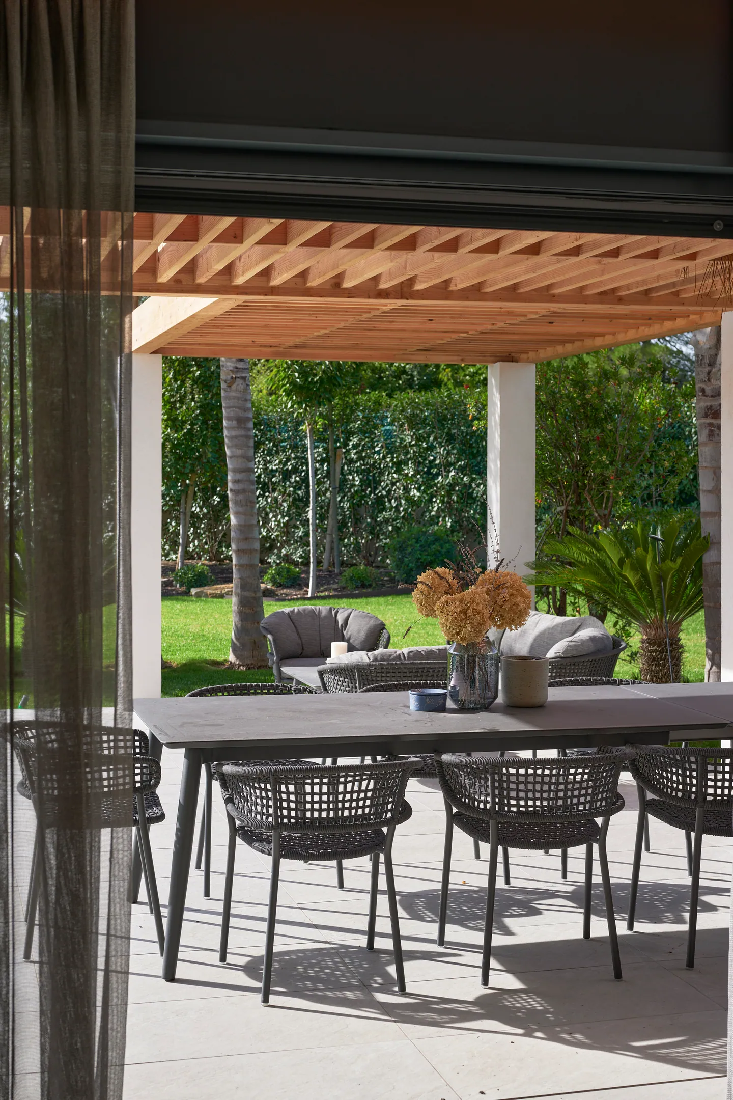 Table de repas extérieure sous pergola en bois, salon de jardin et palmiers en arrière-plan