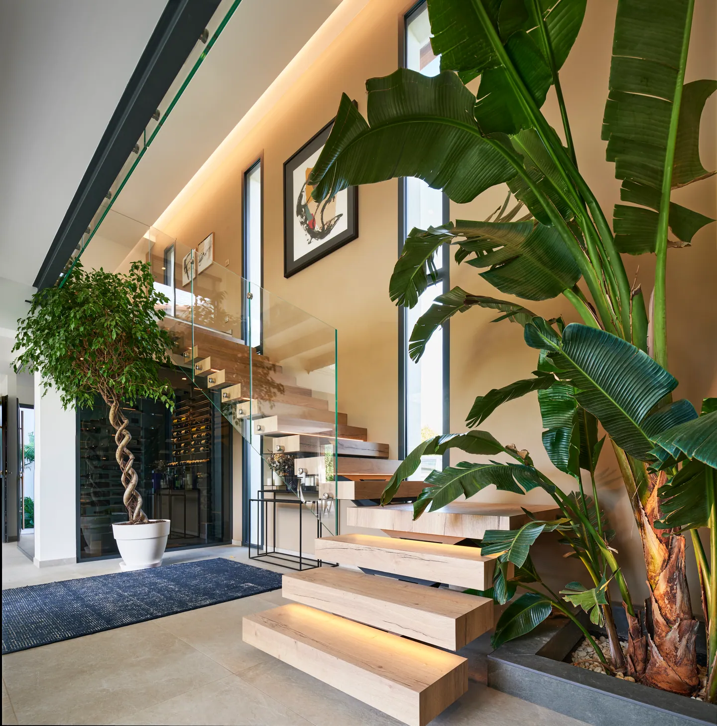 Hall d'escalier aux marches suspendues éclairées, plantes tropicales et cave à vin vitrée