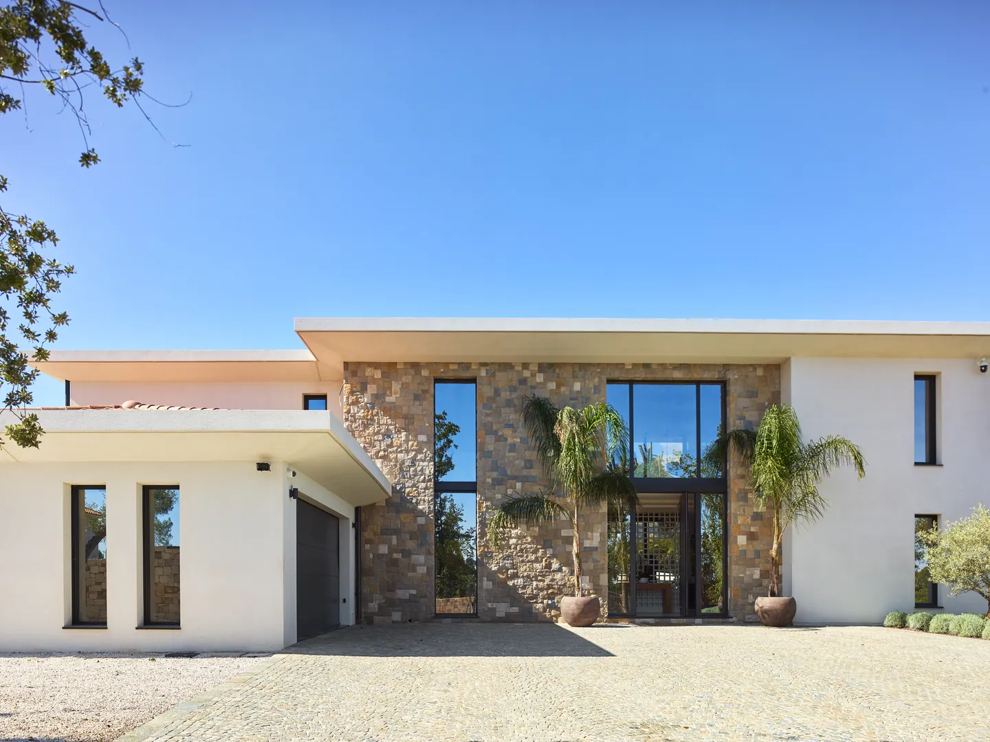 Entrance facade in stone and white render, full-height glazing and potted palms