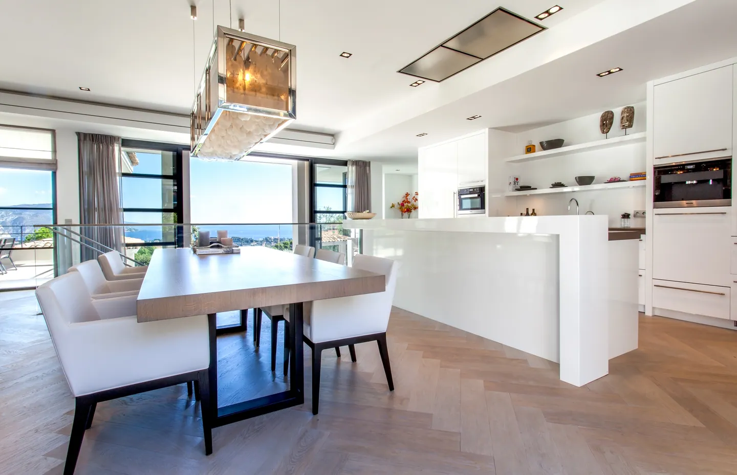 Wooden dining table under a chandelier, white kitchen and windows overlooking the sea