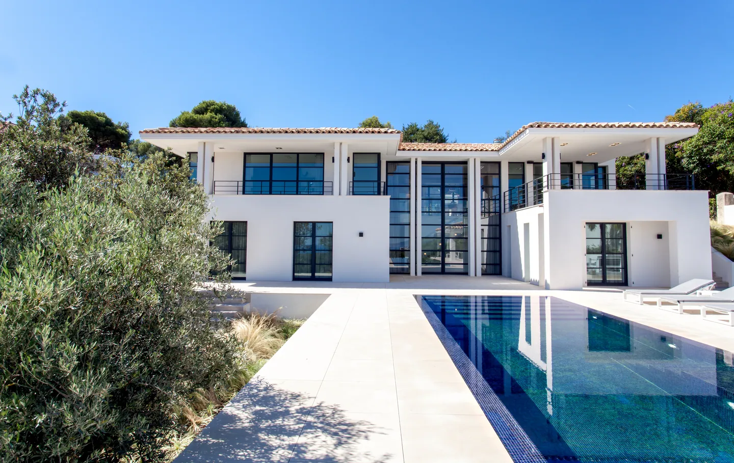 Façade blanche symétrique aux grandes menuiseries noires, piscine à débordement au premier plan