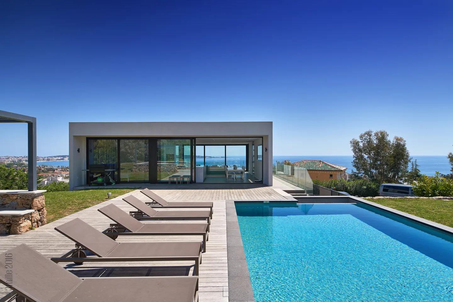 Piscine bordée de transats sur deck en bois, villa vitrée et horizon marin