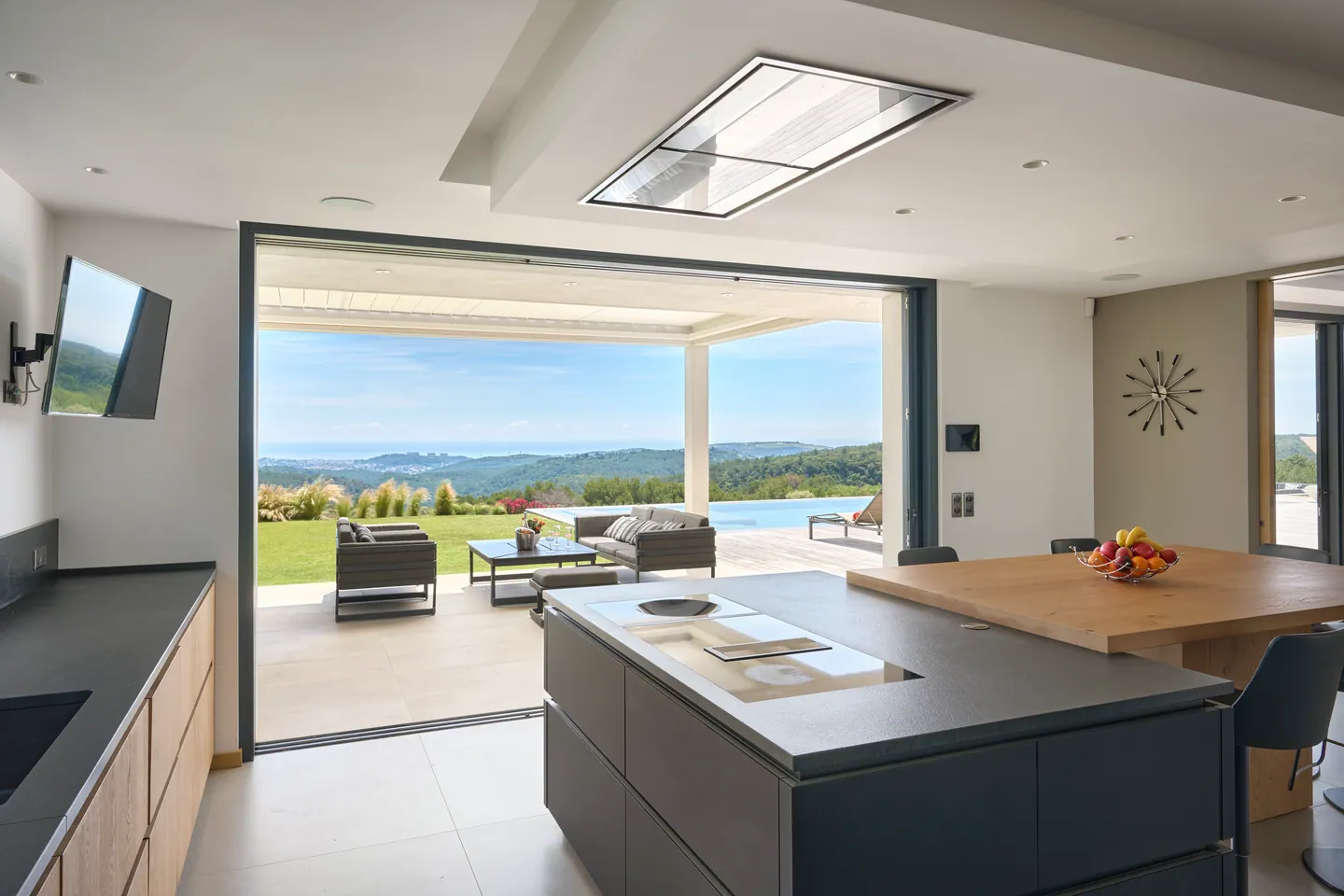 Îlot de cuisine anthracite et table en chêne, baie ouverte sur la terrasse et la piscine