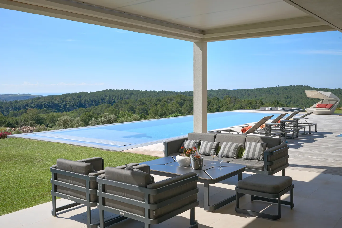 Terrasse couverte avec canapé et fauteuils face à la piscine et aux collines boisées