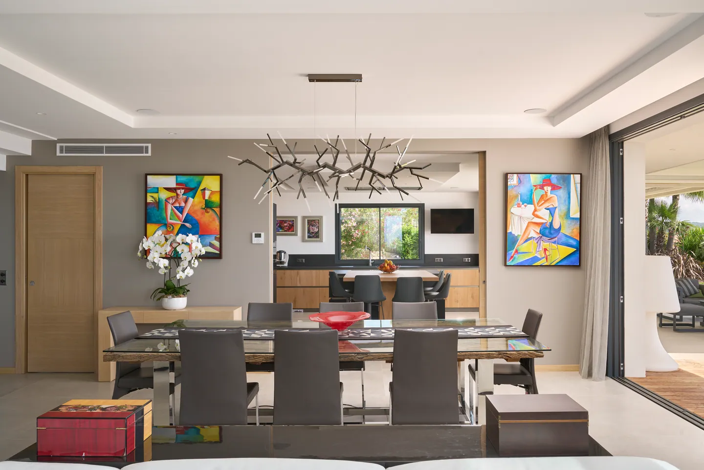 Salle à manger encadrée de tableaux colorés, cuisine visible en arrière-plan