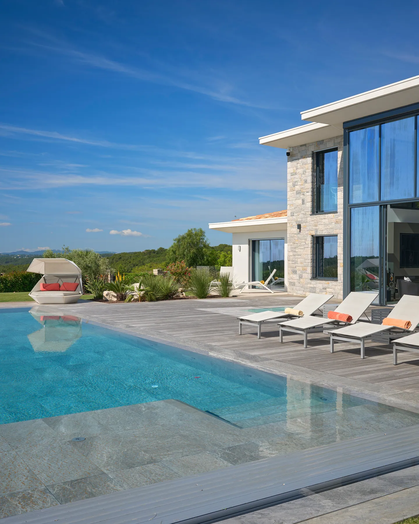 Bord de piscine en pierre, transats et façade vitrée sous ciel bleu