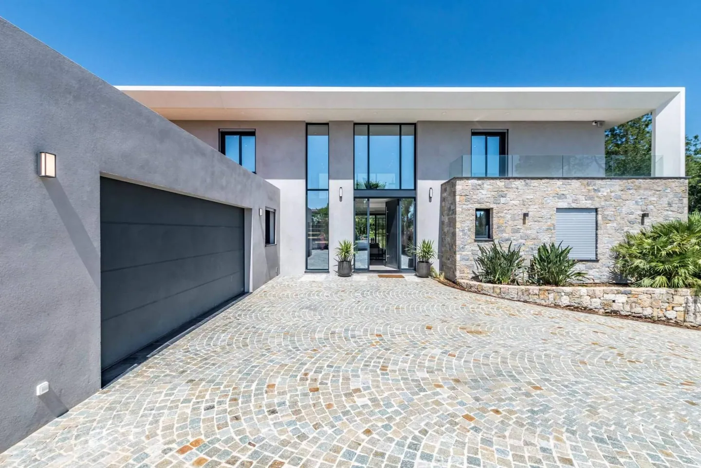 Entrée de la villa : cour pavée en éventail, porte de garage et façade vitrée encadrée de pierre