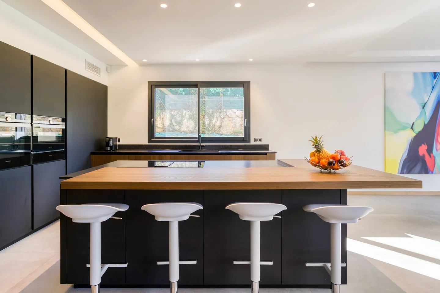 Îlot de cuisine avec plan en bois et quatre tabourets blancs, fenêtre sur un mur de pierre