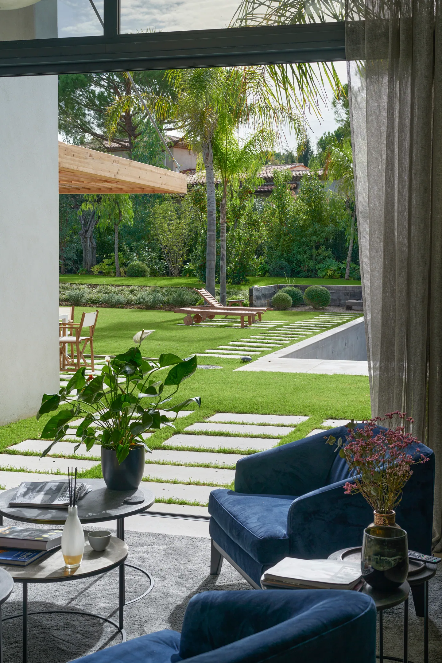 Vue du salon sur le jardin, l'allée dallée et la piscine, fauteuil bleu au premier plan
