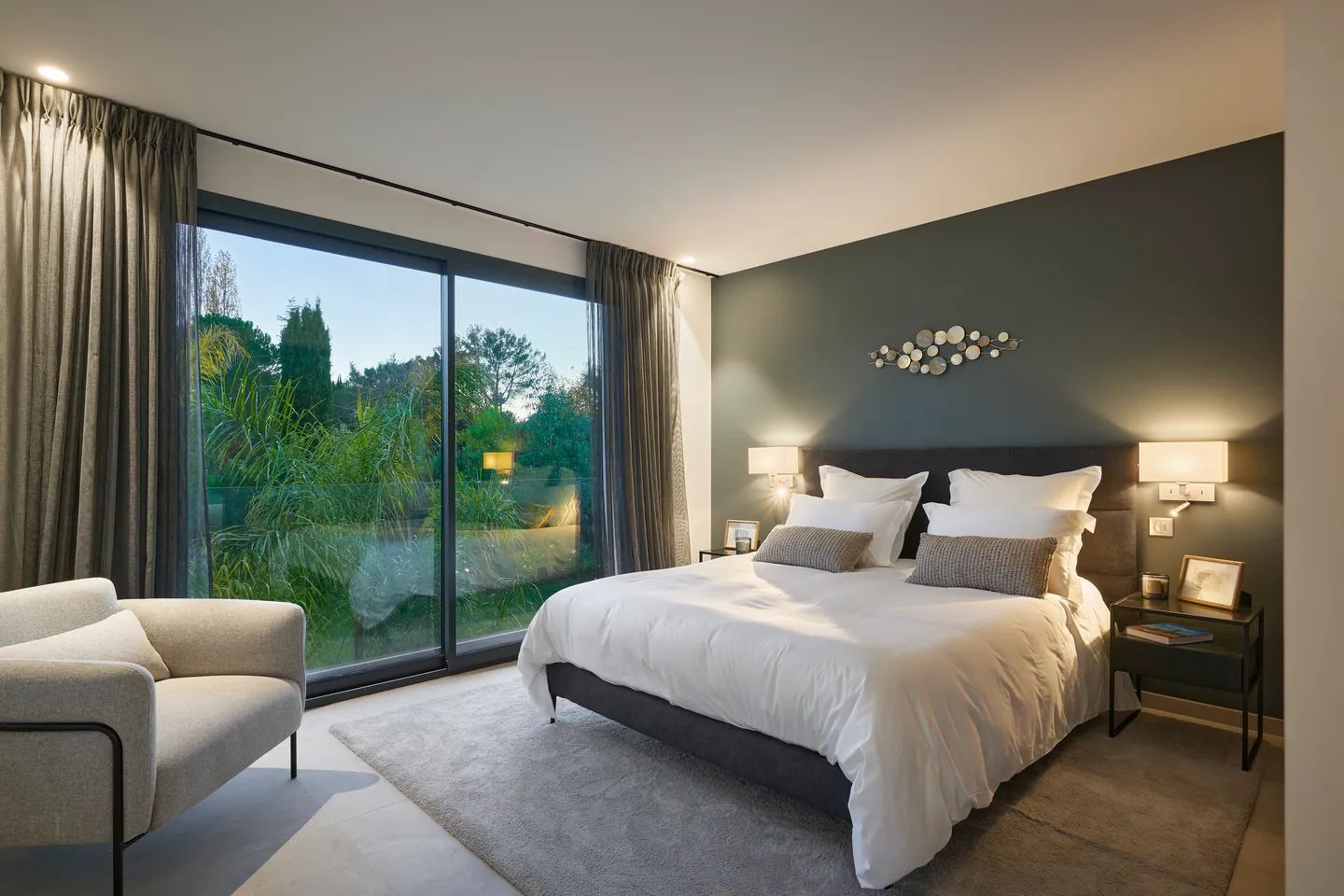 Chambre au crépuscule, lit blanc, mur vert sombre et baie vitrée ouverte sur le jardin