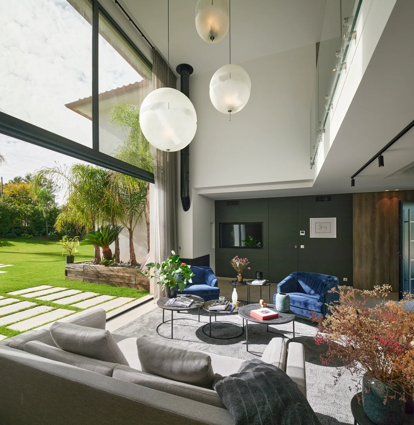 Salon sous verrière double hauteur, globes suspendus et vue sur le jardin et les palmiers
