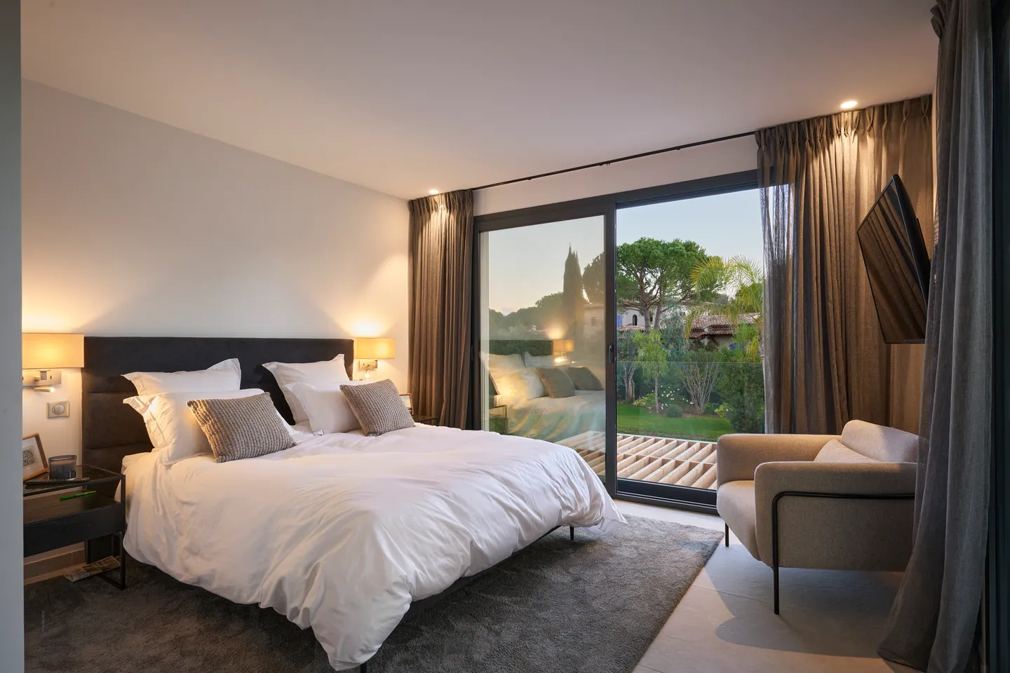 Chambre éclairée par des appliques, lit blanc, fauteuil et baie vitrée sur la terrasse