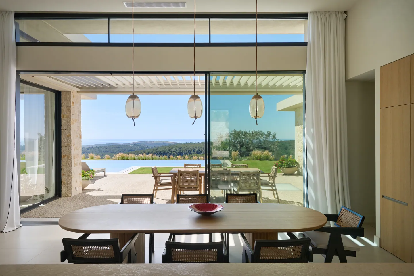 Table à manger ovale face aux baies ouvertes sur la terrasse, la piscine et les collines