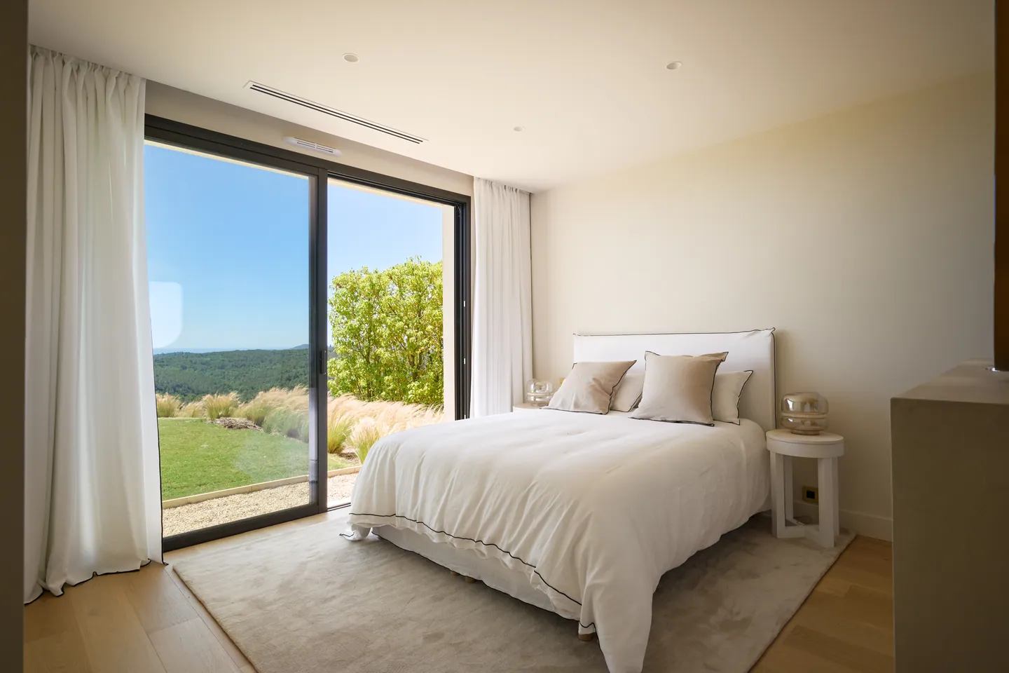 Chambre au lit en lin, grande baie vitrée ouverte sur le jardin et les collines