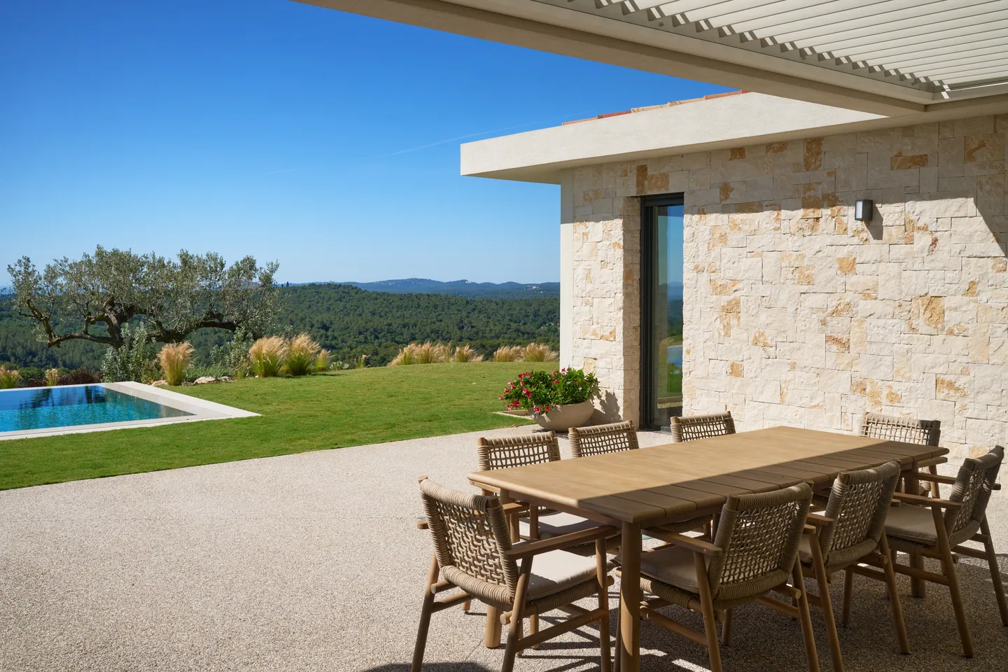 Table à manger extérieure sous pergola, mur en pierre, piscine et olivier en arrière-plan