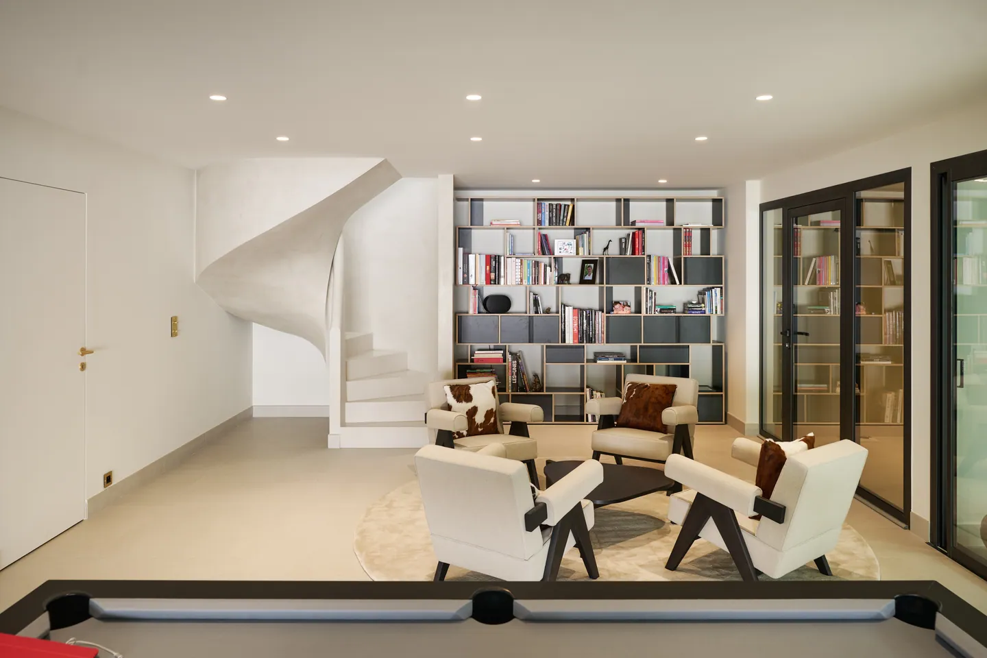 Salon bibliothèque au niveau inférieur, escalier sculpté, fauteuils sur tapis rond et billard