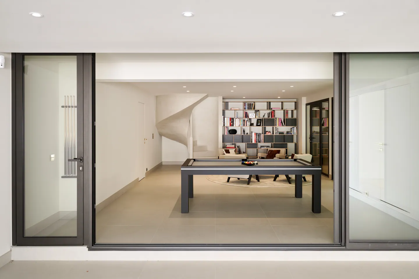 Salle de jeux avec billard, bibliothèque et escalier sculpté vus à travers une baie vitrée