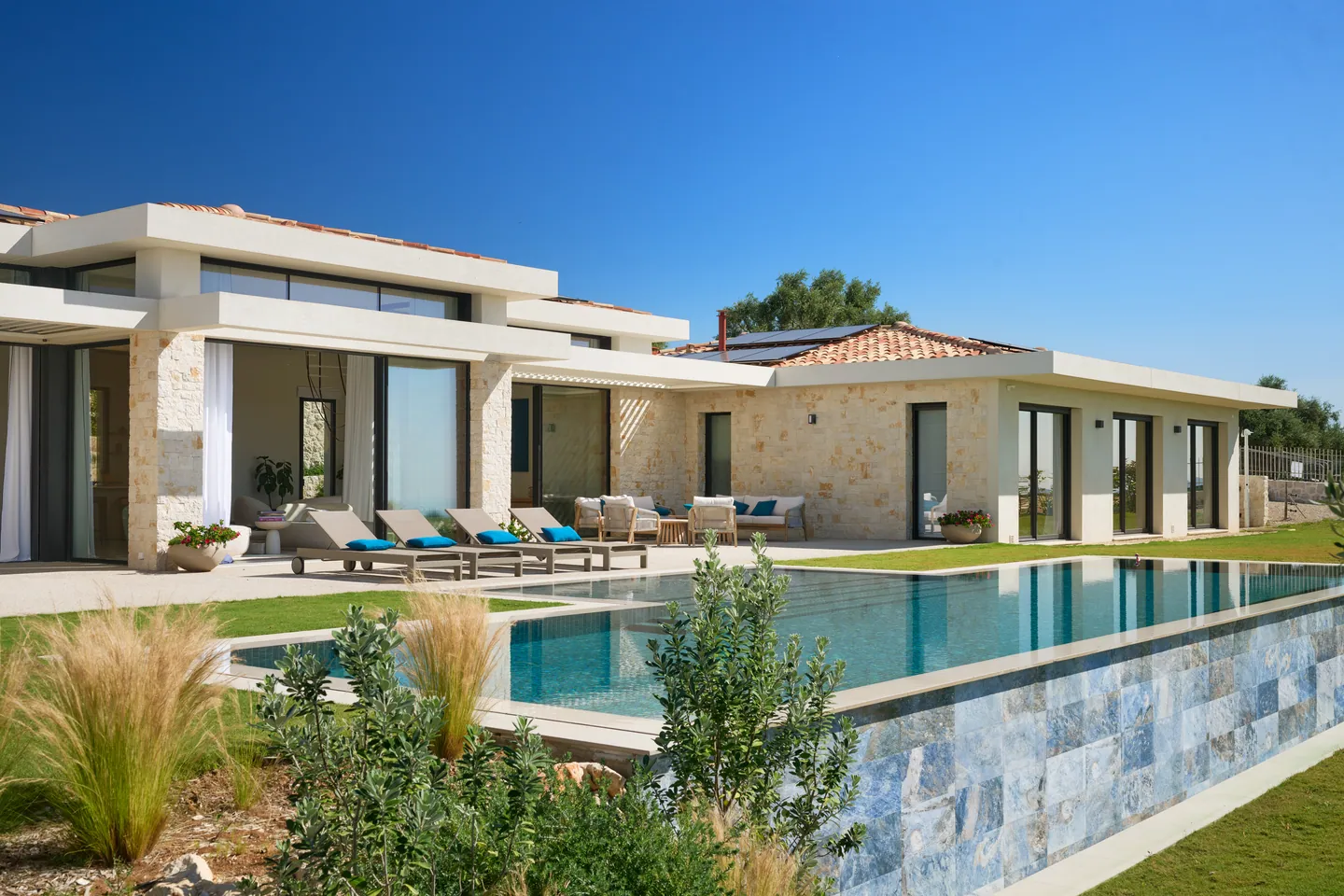 Piscine à débordement habillée de pierre, transats et façade aux grandes baies vitrées