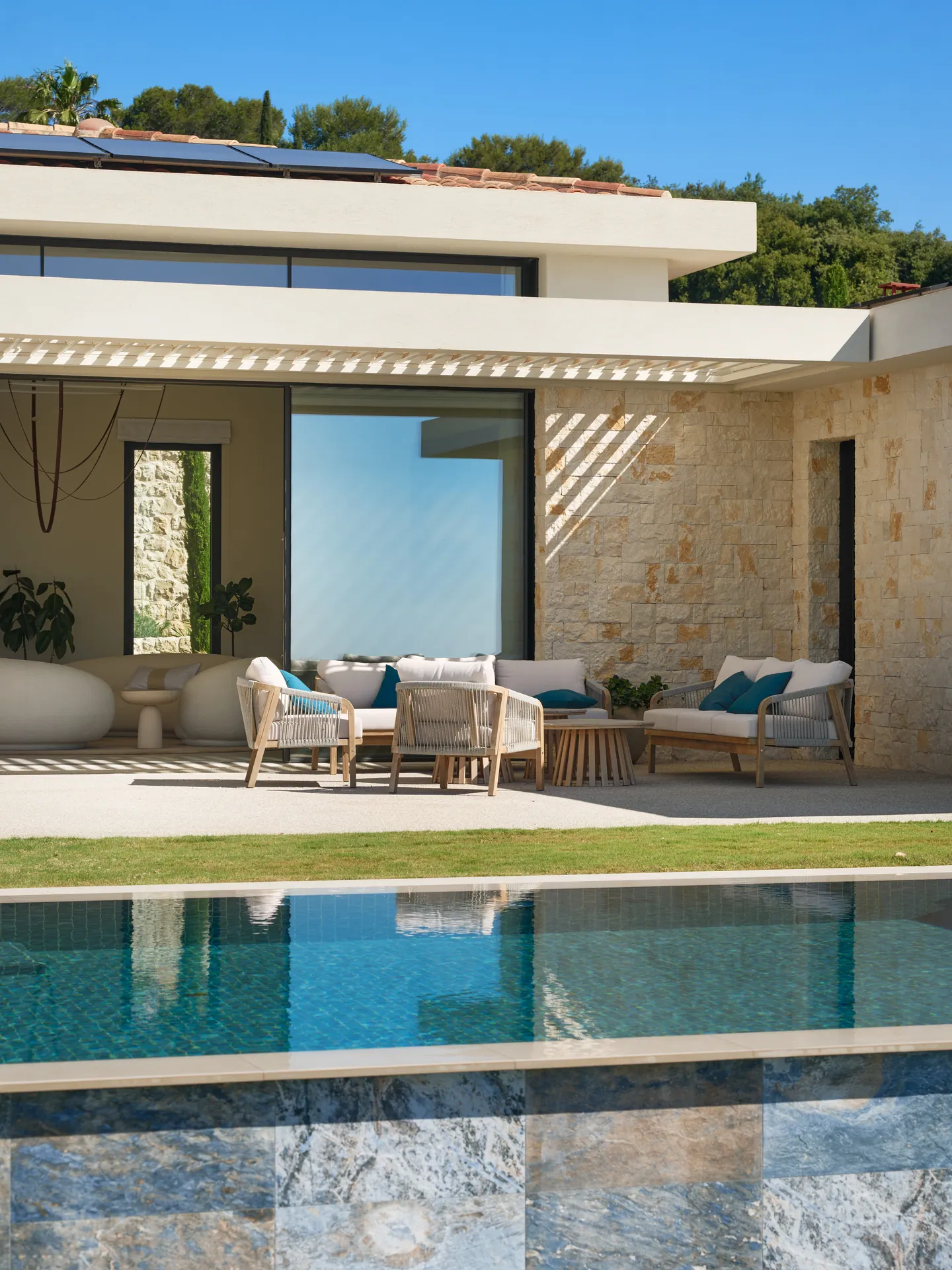 Salon d'extérieur sous pergola bioclimatique, murs en pierre claire, piscine au premier plan