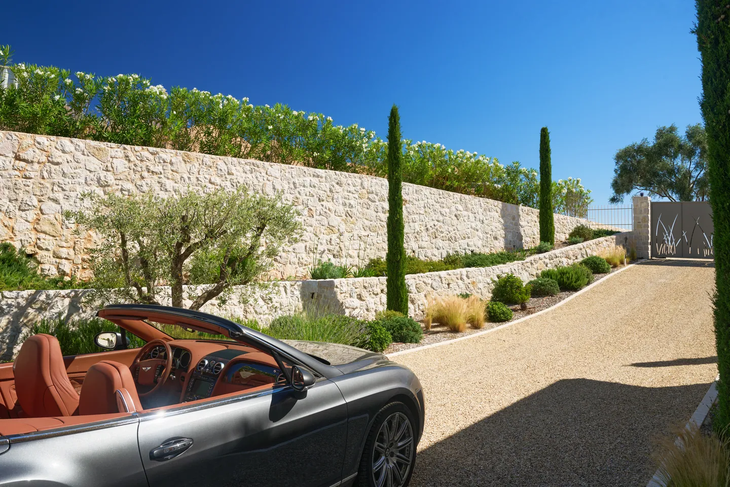Cabriolet garé dans l'allée, murs de soutènement en pierre plantés d'oliviers et de cyprès