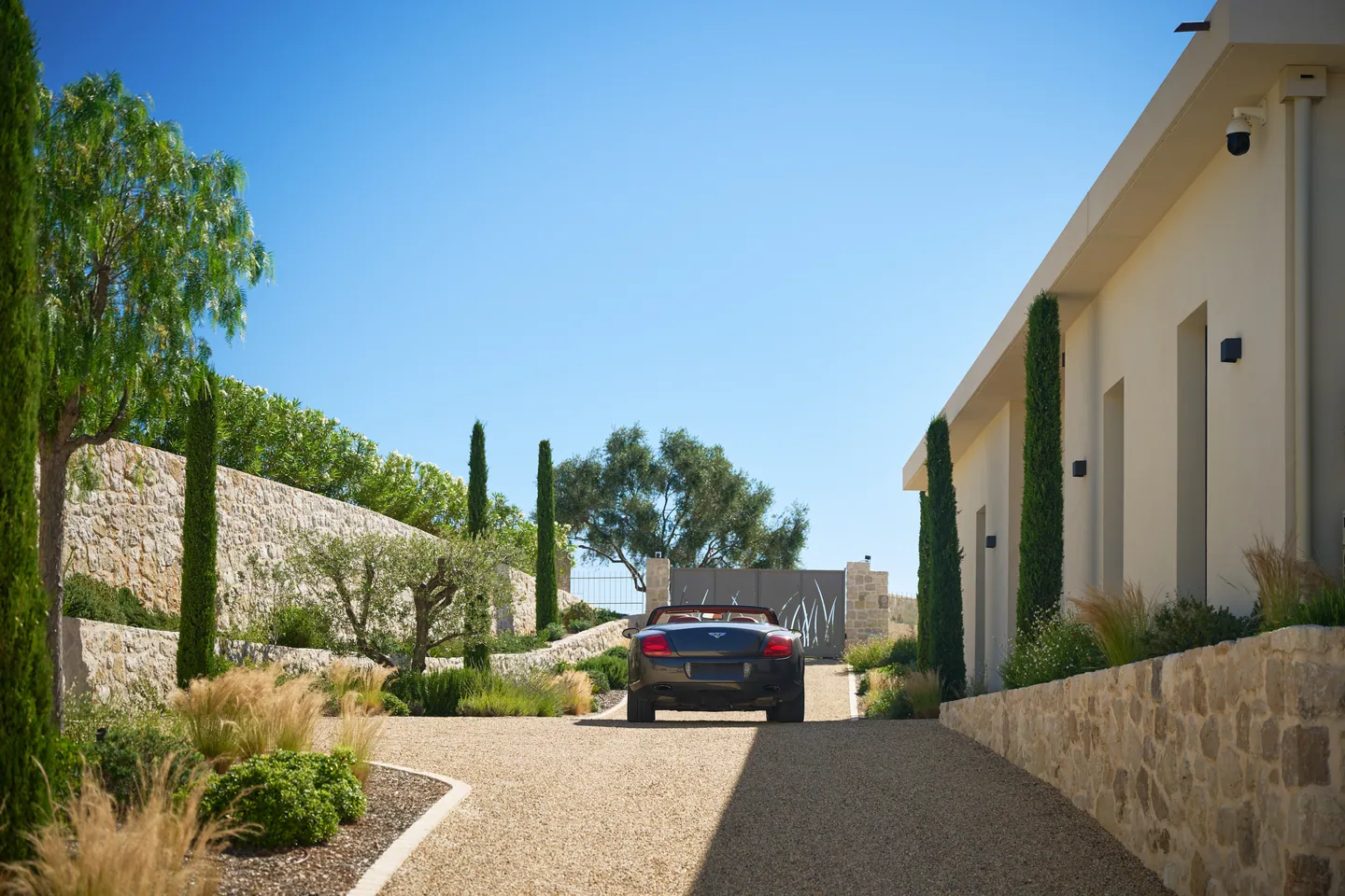 Allée en gravier bordée de cyprès et de murs en pierre, voiture face au portail d'entrée