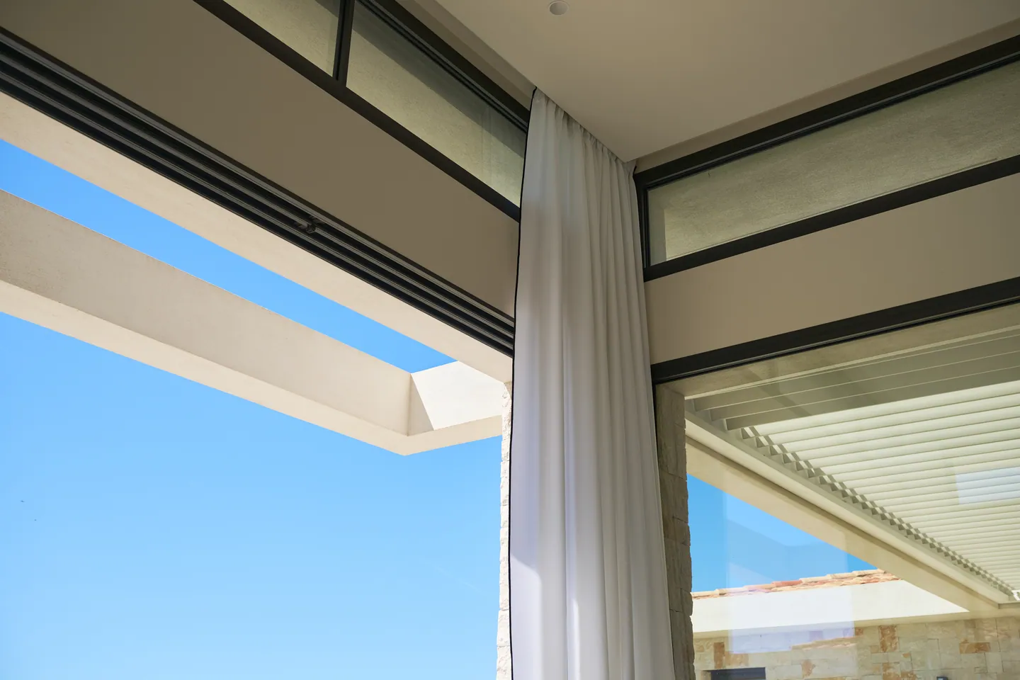 Voilage blanc entre deux baies vitrées à profils noirs, pergola à lames en arrière-plan