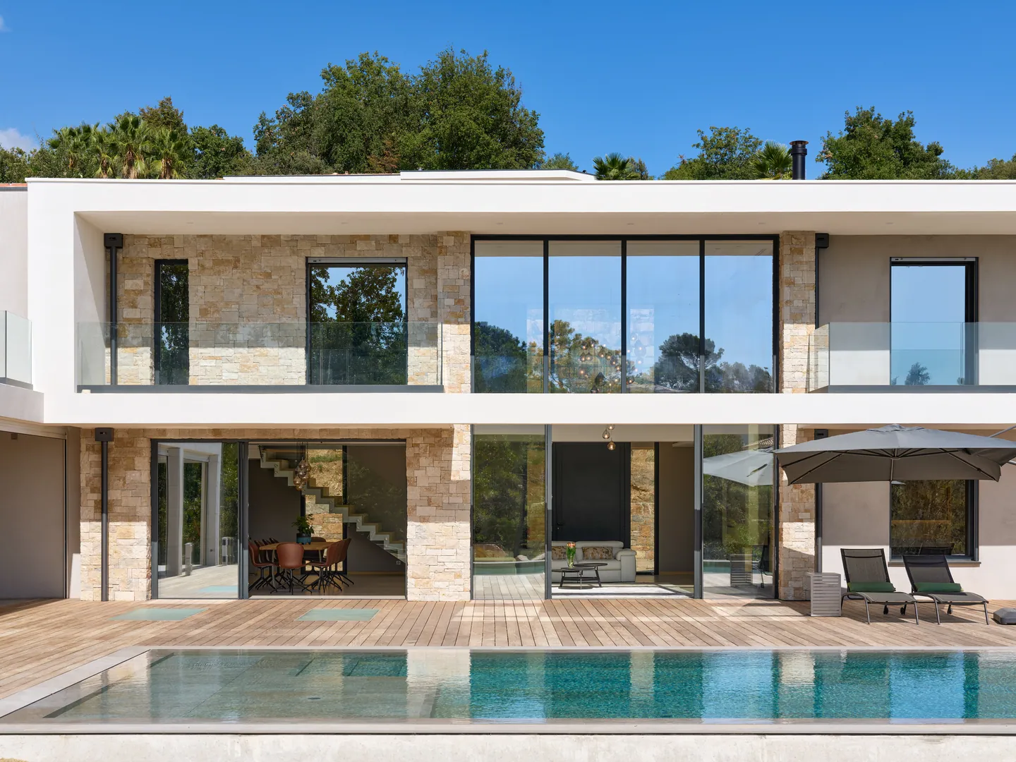 Façade en pierre et baies vitrées toute hauteur, terrasse en bois et piscine au premier plan