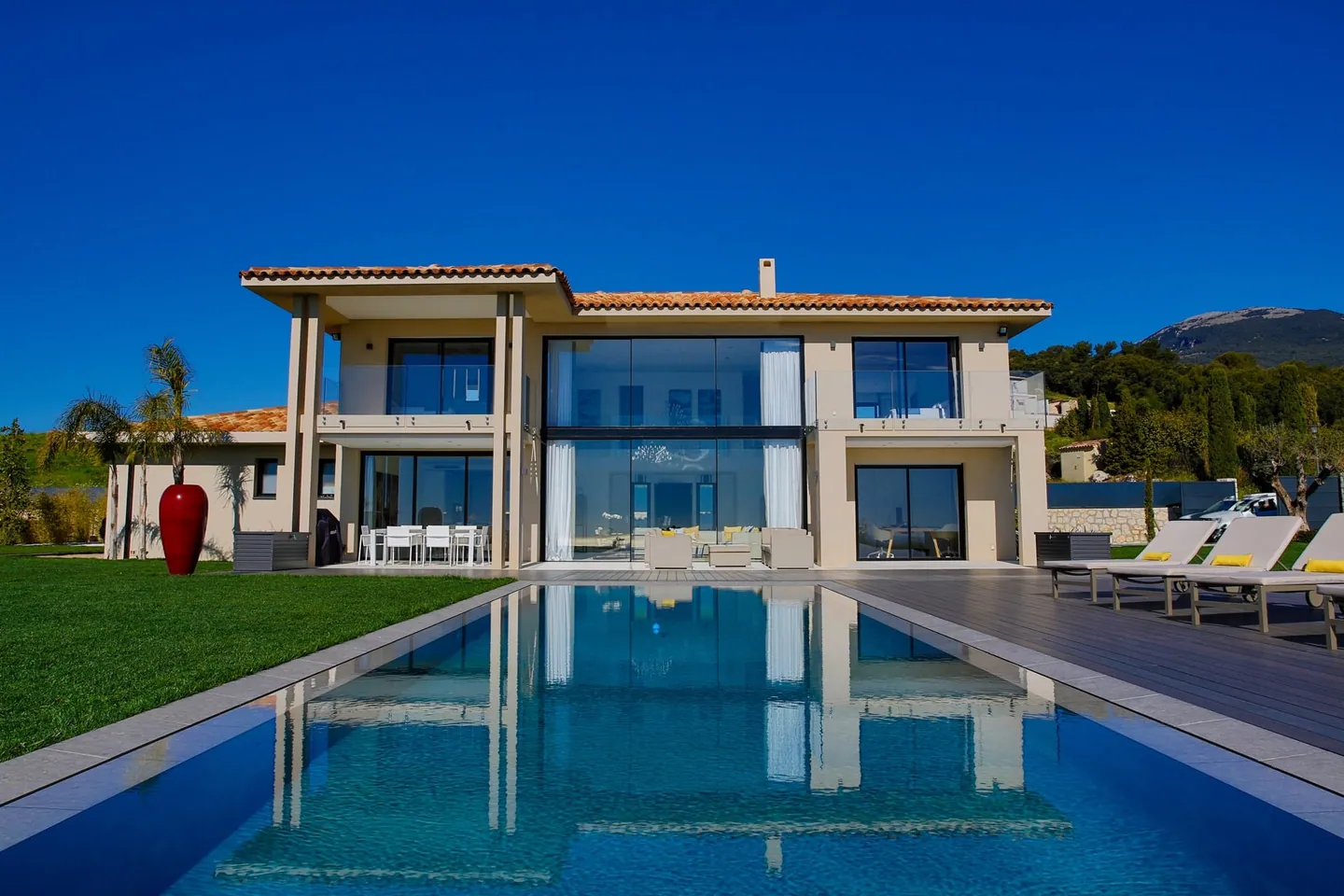 Two-storey villa under tiled roofs reflected in a long pool, with lawn and sun loungers