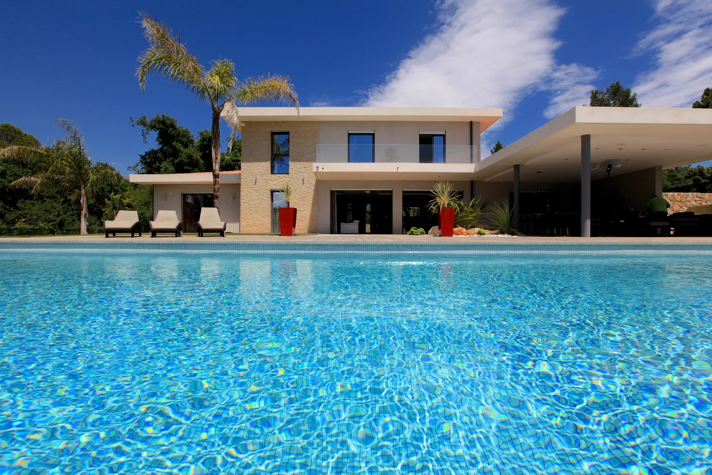 Villa contemporaine en pierre et enduit clair vue depuis la piscine, palmiers et transats